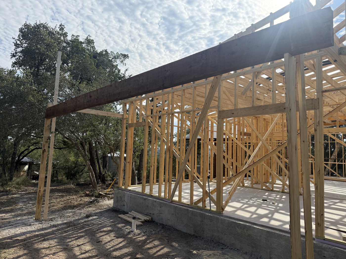 Hill Country Escape, a custom home under construction with a heavy timber porch beam, wood framing and live oaks beyond