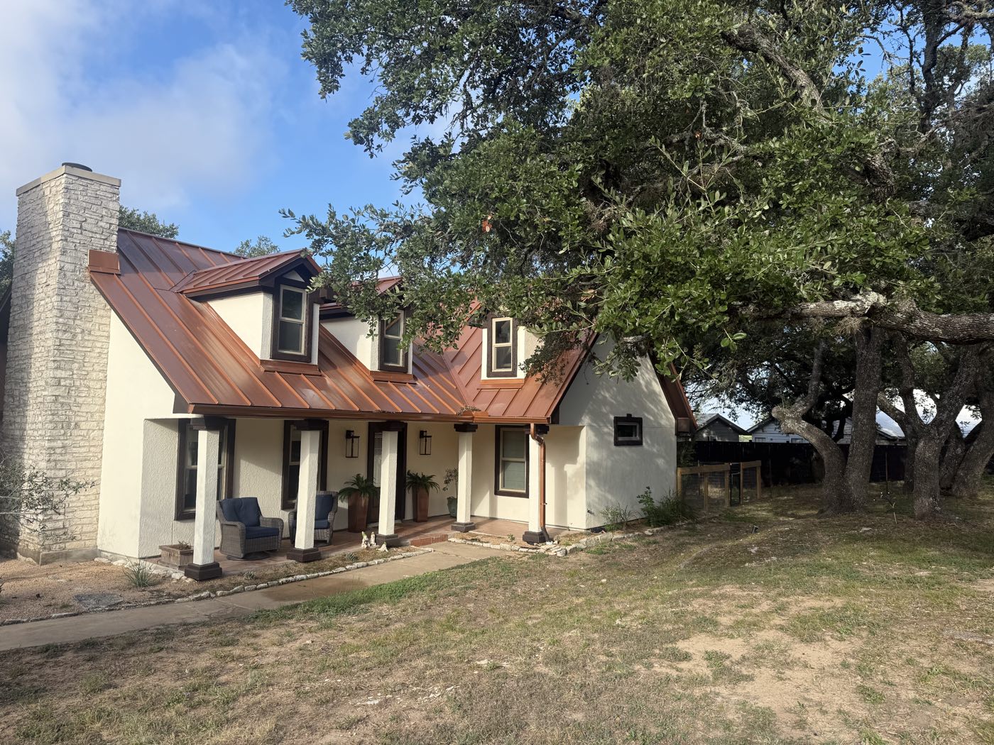 Modern rustic Hill Country home with a standing-seam metal roof, limestone chimney and covered porch, set among live oaks